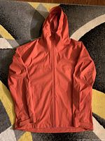 Front view of coral/orange Uniqlo rain jacket laid flat, showing hood, zipper, and sleeves.