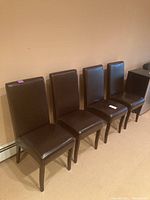 Four brown leather-like dining chairs lined up against a beige wall.