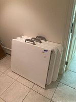 Three white plastic Cosco folding tables stacked with handles visible, stored upright against a wall on a tiled floor.