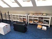 Wide angle photo showing three identical white cubby bookshelf units side by side along a wall under skylights. Each unit is 68x15x29 inches. The shelves hold stacks of brown and manila file folders and papers. A large black case and a cardboard box are near the units on a patterned carpet.