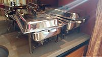 Photo showing two chafing dishes placed side by side reflecting light, with lids on and black handles visible.