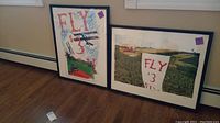 Two framed wall art prints side by side on floor against wall, showing 'FLY $3' text and vintage airplane images.
