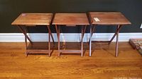 Three wooden snack trays set upright, showing foldable legs and rectangular flat tops, positioned side by side against a dark wall and wooden floor.