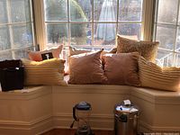 Six decorative pillows arranged on a window seat with a dark brown leather storage cube and two kitchen appliances below the shelf.