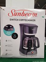 Boxed Sunbeam Switch Coffeemaker showing front with product image and feature details including 12-cup capacity, on/off indicator light, easy-view water window, washable filter basket, and cord storage.