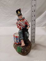 Front view of seated drummer boy figurine beside a measuring ruler showing height about 6 inches.