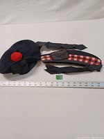 Photo showing two Highland military hats on a white background with a ruler for scale. Left is a navy blue Tam O'Shanter hat with a red pom-pom and black bow. Right is a diced red and white checkered Glengarry hat with attached black ribbons and leather badge holder.