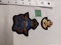 Photo showing two RCAF badges on a white surface with a ruler for scale, one metal braid blazer crest and one cloth hat badge.