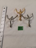 Top view of three military boot spurs: one gold-colored single spur centered with two silver-colored spurs on either side, alongside a measuring tape for scale.