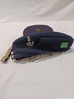 Side view of dark blue wedge cap with brass anchor badge and button detail, laid on side with chin strap visible