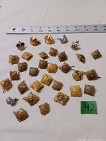 Top-down photo showing 39 assorted Canadian military infantry metal rank pips and crowns arranged in rows with a ruler for scale, varying in gold and silver colors with different backings including wires and hoops.