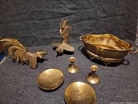 Photo showing two brass rooster figurines, a decorative brass bowl with handles and feet, two small brass candleholders, and two small brass dishes or bowls on dark fabric background.