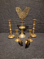 Front view of five brass candlesticks and two brass horse figurines arranged on gray surface with black backdrop.