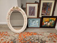 Photo showing an oval white plaster frame with floral decorations, three Asian-inspired framed prints, and one framed watercolor landscape painting arranged on the floor.