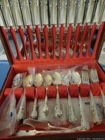 Photo showing the full set of 12 silver plated flatware pieces in original red velvet-lined presentation box with slots for each utensil, including knives, forks, spoons, and serving utensils.