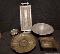 Image showing five decorative pewter vessels on black background: rectangular engraved tray, round bowl with circular indentations on rim, fluted round dish with central emblem, small goblet-shaped cup, square ribbed planter bowl.