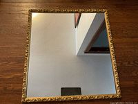 Full front view of the nearly square antique gold gilt framed mirror resting on wooden floor, showing reflective mirror surface and outer decorative frame.