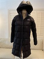 Full front view of the black North Face down-filled coat with hood up, showing length and quilted design.