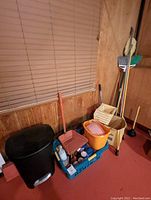 Wide view of the cleaning supplies grouped together against the wood panel wall including buckets, trash can, and cleaning bottles