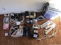 Lot showing various electrical items on carpeted floor next to wall including power strips, two cameras, black tripod with case, alarm clocks, flash light, remote control, cables, and antennas.