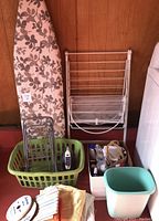 Photo of ironing board with floral cover, drying rack, green laundry basket with Rowenta iron and metal rack parts, and assorted smaller items including folded fabric and ribbons.