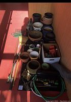 Overview photo of planter pots and garden tools including black plastic pots, terracotta pots, metal rake, broom, garden hose, toolbox with small tools and accessories