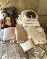 Wide view showing pile of assorted bedding items including pillows, blankets, and comforters stacked on bed and chair in a room with beige walls