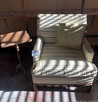 Front view of striped upholstered chair with wood frame and green pillow, along with wooden side table on the left.