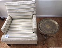 Striped upholstered chair and tarnished brass side table on wooden floor, against white wall.