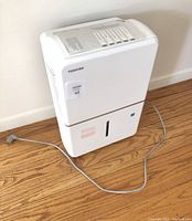 Front view of white Toshiba dehumidifier placed on wooden floor, showing full unit with power cord.