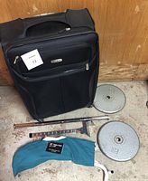 Front view of packed Samsonite suitcase with barbell plate, travel chair fabric sleeve, and metal closet bar visible in front on floor beneath.