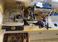 Wide view showing metal dish drying rack, cutlery drawer open with spoons and utensils, assortment of knives including Hamilton Beach electric cutlery knives, scissors, skewers, cooking utensils, wine tools, grill fork, ceramic spoon rest, and metal sharpener all arranged on countertop.