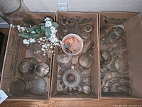 Top view of three cardboard boxes containing glassware including decanters, vases, drinking glasses, glass fish decor, artificial flowers in a vase, and a ceramic bowl.