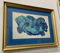 Front angle view of framed watercolor painting with blue mat and gold frame hung on wall.