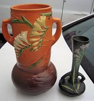 Photo of both vases showing front and side views; tall orange-brown vase with floral relief and two handles, smaller green vase with flared neck