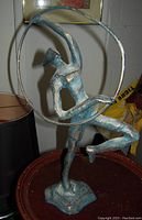 Front angle of bronzed metal statue depicting a female figure holding a hoop, standing on one leg, showing textured metal and patina finish.