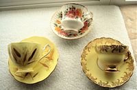 Top angle showing the three porcelain teacup and saucer sets with distinct floral patterns and gold accents on white and yellow backgrounds.