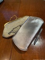Photo of two metallic clutch purses; one gold with a chain and one silver, placed on a wooden surface with a pen for scale.
