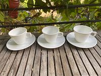Three white Corelle coffee cups each with an accompanying saucer arranged on a wooden surface outdoors, showing the set’s shape and size.