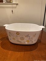 Front view of white rectangular Corning Ware casserole dish with floral pattern in yellow, white and blue on side, placed on wooden table.