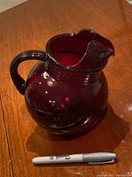 Full view of red glass jug showing the rounded shape, handle and spout with a marker for scale.