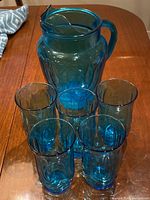 Photo showing one turquoise tall water jug with hinged lid and handle, surrounded by five matching turquoise water glasses arranged in front on a wooden table.