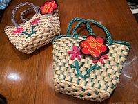 Photo showing two kids raffia baskets on a wooden surface; one smaller beige basket with pink flower decoration and braided purple handle, and one larger beige and teal basket with pink flower decoration and braided teal handle; both with original tags attached.