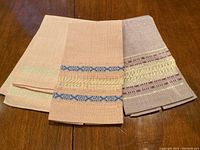 Three handwoven linens arranged overlapping on wooden surface showing decorative woven bands and fine fabric texture