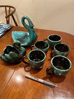 Two Blue Mountain Pottery swan figurines and five matching coffee cups on a wooden table with a Sharpie for scale.