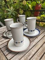 Four tall mid-century modern coffee cups with speckled beige finish and blue/tan dot pattern, placed on wooden table outdoors with greenery background