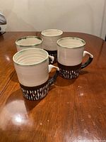 Four handmade mid-century modern mugs on wooden table, showing white glazed mug with green rim and brown patterned band near base.