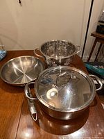 Photo showing all four items: saucepan with lid on, frying pan, and colander. Items placed on wood table.