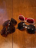 Three pairs of sunglasses placed on a wooden surface: black round frame, tortoiseshell frame, white frame with red temples and lenses.