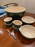 Large and medium green and cream Emile Henry ceramic bowls and small ramekins arranged on wood table with marker for scale.